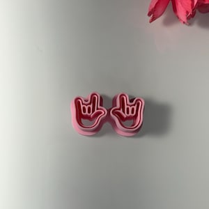 May include: A light pink plastic cookie cutter shaped like the 'I love you' hand gesture. The cutter shows the hand sign in detail. The background is white with a pink flower in the top right corner.