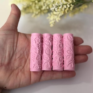May include: Four pink cylindrical rollers with embossed botanical designs, including grapes, strawberries, and leaves. The rollers are held in a hand, with a blurred background of yellow flowers.