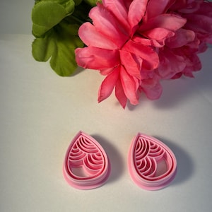 May include: Two pink teardrop-shaped cookie cutters with an intricate interior design. The cutters are on a white surface, with pink flowers and green leaves in the background. The cutters are likely for making decorative cookies.