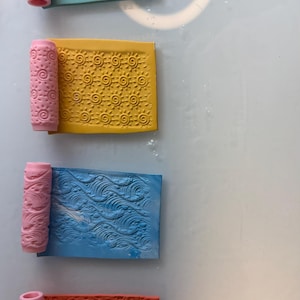 May include: Four colorful clay texture rollers and matching rectangular clay pieces. The rollers and clay are pink, teal, yellow, blue, and red. The clay pieces have embossed patterns of leaves, suns, waves, and flowers. The rollers are cylindrical.