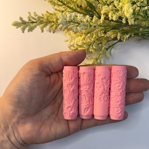 May include: Four pink cylindrical rollers with floral patterns are held in a hand. The rollers have detailed embossed designs of flowers and leaves. The background includes yellow and green foliage.