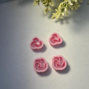 May include: Four small, pink, flower-shaped cookie cutters. Each cutter has a three-petal design with a circular center. The cutters are arranged on a white surface, with yellow and white flowers in the background.