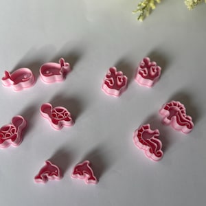 May include: A collection of pink plastic cookie cutters in various sea creature shapes, including a whale, turtle, dolphin, seahorse, and coral. These cutters are designed for creating ocean-themed treats.