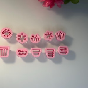 May include: A collection of pink plastic cookie cutters in various floral and geometric shapes. The set includes flower, sunburst, and basket designs. Ideal for baking and crafting projects. The cutters are arranged on a white surface.