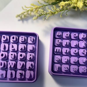 May include: Two purple alphabet stamp sets. Each set is in a square tray with individual letter stamps. The letters are in a sans-serif font. The trays are on a white surface with a sprig of yellow flowers.