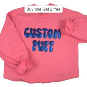 Pode incluir: Uma camisola coral com mangas compridas. As palavras "CUSTOM PUFF" são bordadas em letras azuis com bolhas na frente. Acima do texto há um retângulo branco com o texto "Buy one Get 2 free".