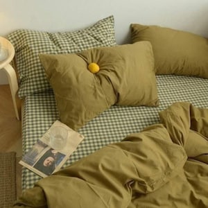 May include: A bed with olive green bedding and pillows, including a decorative pillow with a yellow button. The bedspread and pillowcases feature a green and white checkered pattern. A newspaper and glass are on the bed.