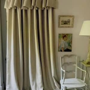 May include: Cream-coloured striped curtains with a valance, suspended from a black rod. The curtains feature vertical stripes in a darker neutral hue. The image also shows a white chair, artwork, and a lamp.