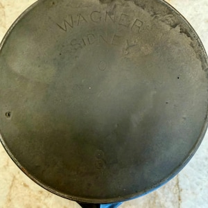 May include: A round, dark gray cast iron skillet base featuring the embossed text WAGNER SIDNEY O and the number 8 centered on the bottom surface. The metal shows a weathered, matte finish with visible signs of use and a circular rim edge.