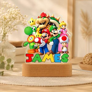 May include: A clear acrylic night light featuring a colorful graphic of Mario, Luigi, Princess Peach, Bowser, Yoshi, Toad, and a star. The name JAMES is printed in large, multi-colored block letters at the base, set on a light wood stand.