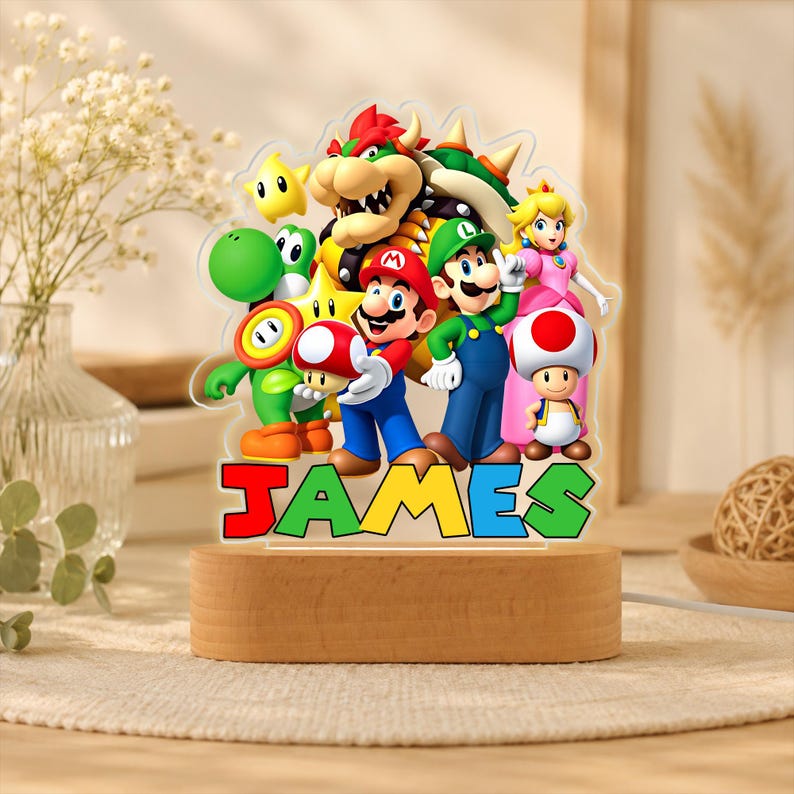 May include: A clear acrylic night light featuring a colorful graphic of Mario, Luigi, Princess Peach, Bowser, Yoshi, Toad, and a star. The name JAMES is printed in large, multi-colored block letters at the base, set on a light wood stand.