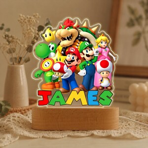 May include: A custom LED night light featuring a colorful acrylic cutout of Mario, Luigi, Princess Peach, Bowser, Yoshi, Toad, and a star. The base is made of light-colored wood. The name JAMES is printed in large, multi-colored block letters at the bottom.