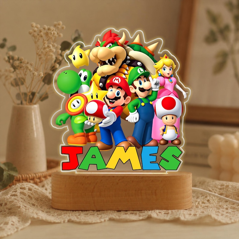 May include: A custom LED night light featuring a colorful acrylic cutout of Mario, Luigi, Princess Peach, Bowser, Yoshi, Toad, and a star. The base is made of light-colored wood. The name JAMES is printed in large, multi-colored block letters at the bottom.