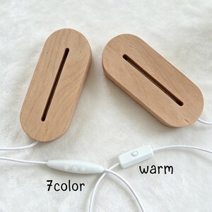 May include: Two oval-shaped light-colored wooden display bases with a central slot for inserts. Each base features a white power cord. One cord includes a controller with three buttons labeled 7color, and the other has a switch labeled warm.