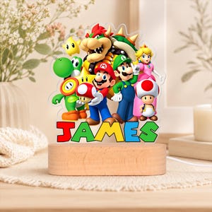 May include: A custom acrylic night light featuring a colorful graphic of Mario, Luigi, Princess Peach, Bowser, Yoshi, and Toad. The name JAMES is printed in bold, multi-colored block letters at the base, which is set into a light wood stand.