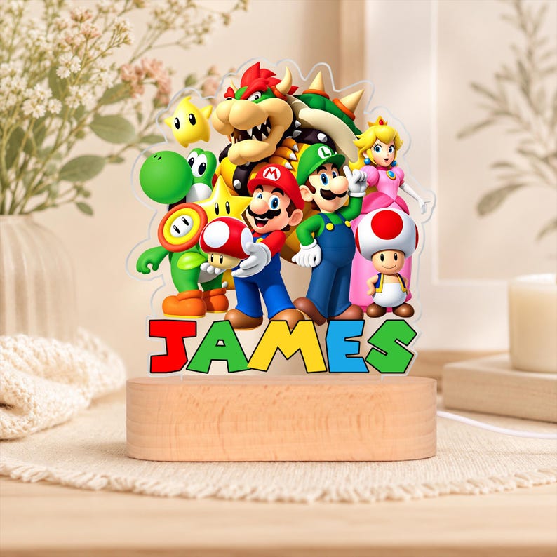 May include: A custom acrylic night light featuring a colorful graphic of Mario, Luigi, Princess Peach, Bowser, Yoshi, and Toad. The name JAMES is printed in bold, multi-colored block letters at the base, which is set into a light wood stand.