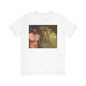 May include: White t-shirt featuring a meme with a shirtless man in sunglasses and a military figure. The meme includes the text "A correr que ahi viene 'El Cuco'" in a speech bubble. The shirt is a casual wear item.
