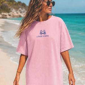 May include: A light pink short-sleeve t-shirt featuring a small blue graphic of a crab centered on the chest. Below the crab illustration, the text reads "a little crabby" in a simple blue font. The shirt has a crew neckline and a loose fit.