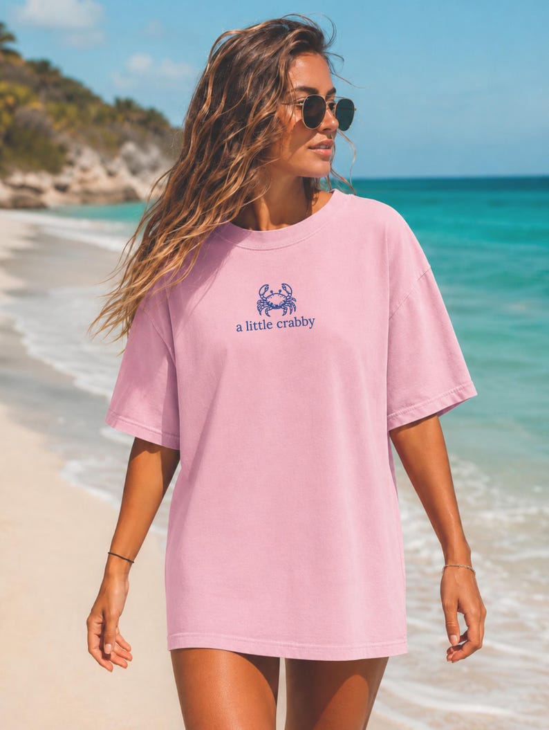 May include: A light pink short-sleeve t-shirt featuring a small blue graphic of a crab centered on the chest. Below the crab illustration, the text reads "a little crabby" in a simple blue font. The shirt has a crew neckline and a loose fit.