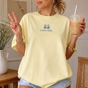 May include: A pale yellow short-sleeve t-shirt featuring a small blue graphic of a crab centered on the chest. Below the crab illustration, the text reads "a little crabby" in a simple blue font. The shirt has a crew neckline and a loose fit.
