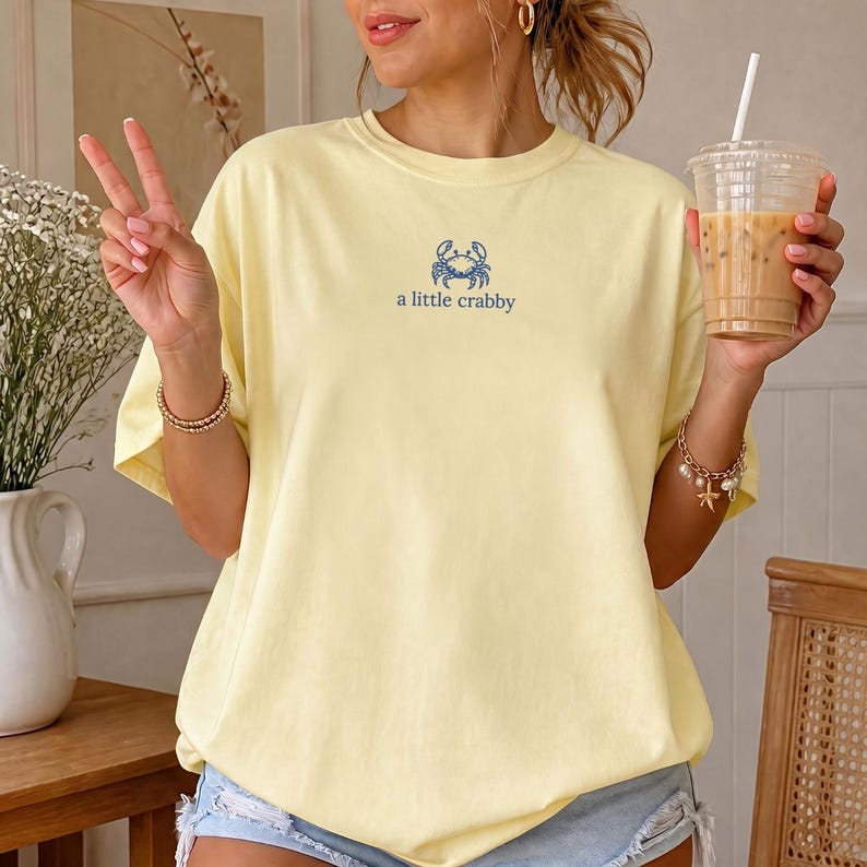 May include: A pale yellow short-sleeve t-shirt featuring a small blue graphic of a crab centered on the chest. Below the crab illustration, the text reads "a little crabby" in a simple blue font. The shirt has a crew neckline and a loose fit.