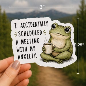Op de afbeelding: Een gestanste sticker met een groene kikker die in kleermakerszit zit en een witte mok vasthoudt. De tekst luidt, I ACCIDENTALLY SCHEDULED A MEETING WITH MY ANXIETY. De sticker is 7,62 cm breed en 5,72 cm hoog.