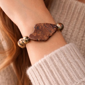 May include: A brown stone bracelet with a large, irregularly shaped central bead. The bracelet features smaller, round beads in shades of brown, green, and gold. The jewelry is worn on a wrist, with a neutral-toned sweater in the background.