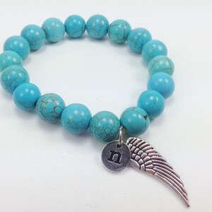 Può includere: Braccialetto di perline blu turchese con un ciondolo d'argento a forma di ala e la lettera "n".