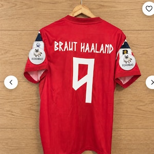 May include: A red short-sleeved sports jersey featuring the white text BRAUT HAALAND and the number 9 on the back. Each sleeve has a circular white patch displaying a cat illustration and the text ECHOMAO, with dark blue and white accents.