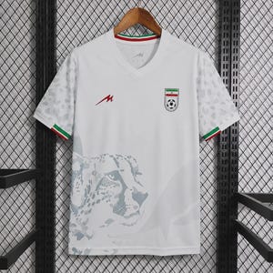 May include: A white short-sleeved V-neck jersey featuring a subtle grey leopard print on the sleeves and lower front. The chest displays a red logo and a crest with the Iranian flag and a soccer ball. The sleeve cuffs have green, white, and red trim.