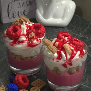 Op de afbeelding: Twee dessertkaarsen in heldere glazen, met lagen roze, witte en bruine was, gegarneerd met rode frambozen en koekjesvormige wasaccenten. Wassen blauwe bessen, wafels en pretzels zijn verspreid. Een bordje leest: "Het geheime ingrediënt is altijd liefde."