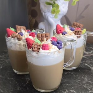 Op de afbeelding: Drie heldere glazen mokken gevuld met een lichtbruine vloeistof, gegarneerd met slagroom en versierd met aardbeien, bosbessen, frambozen en stukjes chocolade. De mokken staan op een donker, patroonrijk oppervlak.