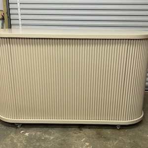 May include: A cream-colored oval-shaped console table or bar unit featuring a vertical fluted or tambour-style exterior design. The piece has a smooth flat top surface and is supported by small casters at the base for mobility.