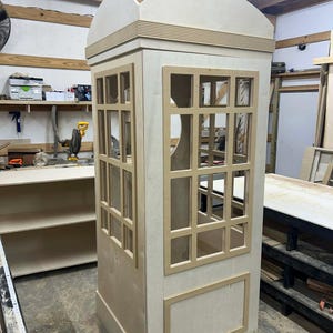 Puede incluir: Una cabina telefónica de madera sin terminar, con una parte superior curvada y ventanas rectangulares. La cabina es de color madera clara y está construida con juntas y molduras visibles. Las ventanas están divididas en múltiples paneles.