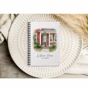 Puede incluir: Un cuaderno con espiral con una ilustración en acuarela de una casa roja con una puerta azul y un perro en el porche. El texto "Welcome Home" y "123 Main St." están escritos debajo de la imagen. El cuaderno está sobre una bandeja decorativa blanca.