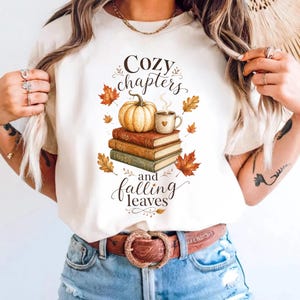 May include: An ivory t-shirt featuring a graphic of three stacked books, a pumpkin, and a steaming mug. The text reads "Cozy chapters and falling leaves" in a decorative script font, surrounded by colorful autumn leaves in shades of orange and brown.