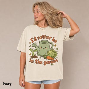 May include: An ivory short-sleeve t-shirt featuring a graphic print of a green watering can with a face, a small sprout, a head of lettuce, and a carrot. The text "I'd rather be in the garden" is printed in brown letters around the illustration.