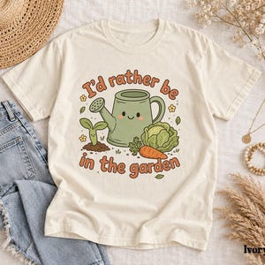 May include: An ivory t-shirt featuring a graphic of a green watering can with a smiling face, a small sprout, a head of lettuce, and a carrot. The text reads, I'd rather be in the garden, in a rounded orange font. The design includes small yellow flowers.