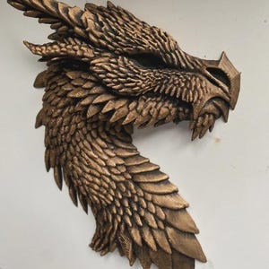May include: A bronze dragon head sculpture with detailed features. The dragon's head is angled, displaying textured scales, sharp teeth, and a detailed horn. The sculpture is mounted on a white surface.
