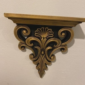 May include: An ornate wall shelf in gold with black detailing. The shelf has a decorative scroll design and a flat top for displaying items. It is mounted on a white wall.