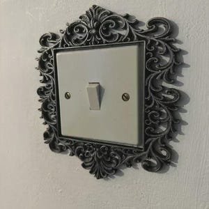 May include: A decorative light switch with a white switch plate and a dark grey ornate frame. The frame features intricate scrollwork and is mounted on a white wall. The switch is in the 'on' position.
