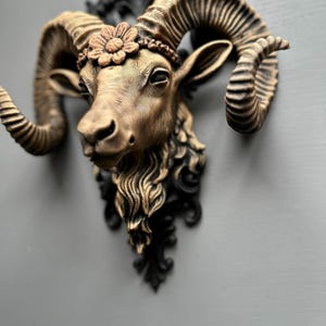 May include: A bronze-coloured ram's head wall sculpture with large, curved horns. The ram wears a flower crown and has a black decorative base. The sculpture is mounted on a grey surface.