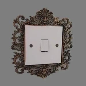 May include: An ornate light switch cover with a bronze-coloured frame. The frame surrounds a white switch plate with a single toggle switch. The design features swirling, floral patterns, adding a vintage aesthetic.