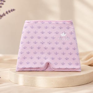 May include: A light purple eye mask with a repeating floral pattern and the text "Serenity". The mask is displayed on a round, light-colored pedestal. Lavender sprigs and a white vase are in the background.