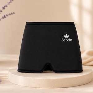 May include: A black eye mask with the brand name "Serein" printed in white. The mask is displayed on a round, light-colored pedestal. A vase of lavender and a lit candle are in the background.