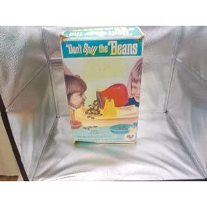 Peut inclure: Une boîte de jeu de société Don't Spill the Beans illustrée avec deux enfants jouant avec un pot rouge et une balance jaune. L'emballage présente le titre du jeu en texte blanc et vert, ainsi que des illustrations de haricots bruns et de bols bleus.