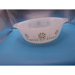 May include: A white oval-shaped glass casserole dish featuring a repeating pattern of olive green flowers and delicate leafy vines around the exterior. The dish has integrated handles on both sides and a smooth, glossy finish throughout the surface.
