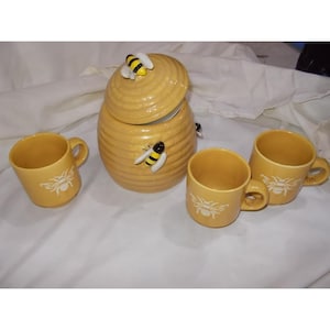 Pode incluir: Um pote de mel de cerâmica amarela em forma de colmeia com tampa, apresentando uma pequena figura de abelha no topo e outra na lateral. O conjunto inclui três canecas amarelas correspondentes, cada uma decorada com uma ilustração de abelha.
