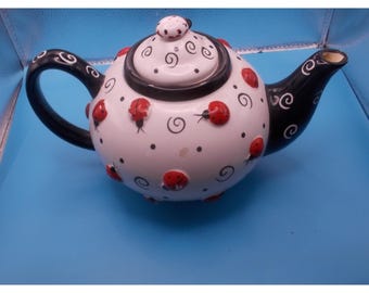 May include: A white ceramic teapot featuring a black handle and spout, decorated with raised red ladybug figures, black polka dots, and black swirling line patterns. The matching lid has a small ladybug knob on top and black swirl details.