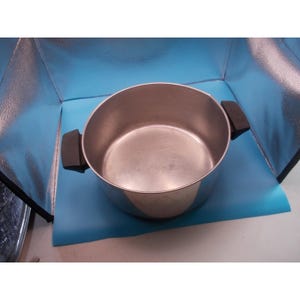 Puede incluir: Una olla de cocina redonda de acero inoxidable de color plateado con una superficie interior lisa. La olla cuenta con dos asas negras de forma rectangular unidas a los lados opuestos del borde exterior para un agarre seguro.